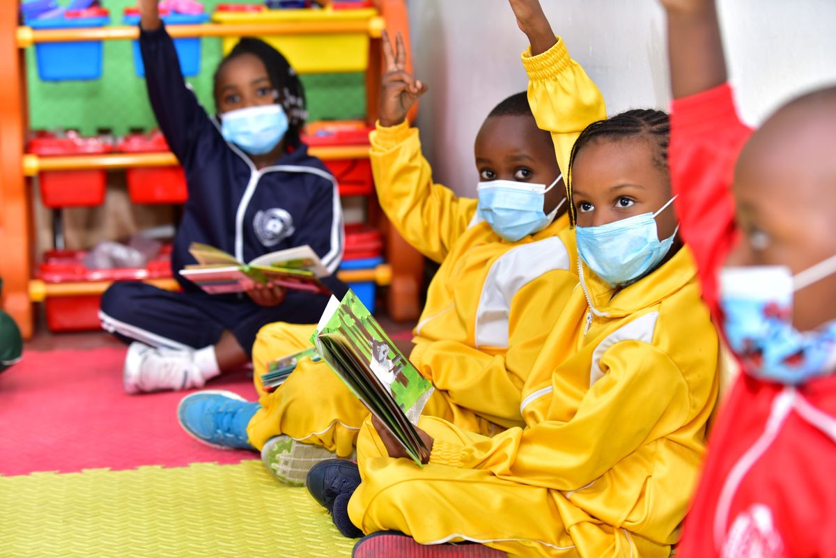 Kindergarten children in yellow tracksuits sitting on the mat with picture books, hands up