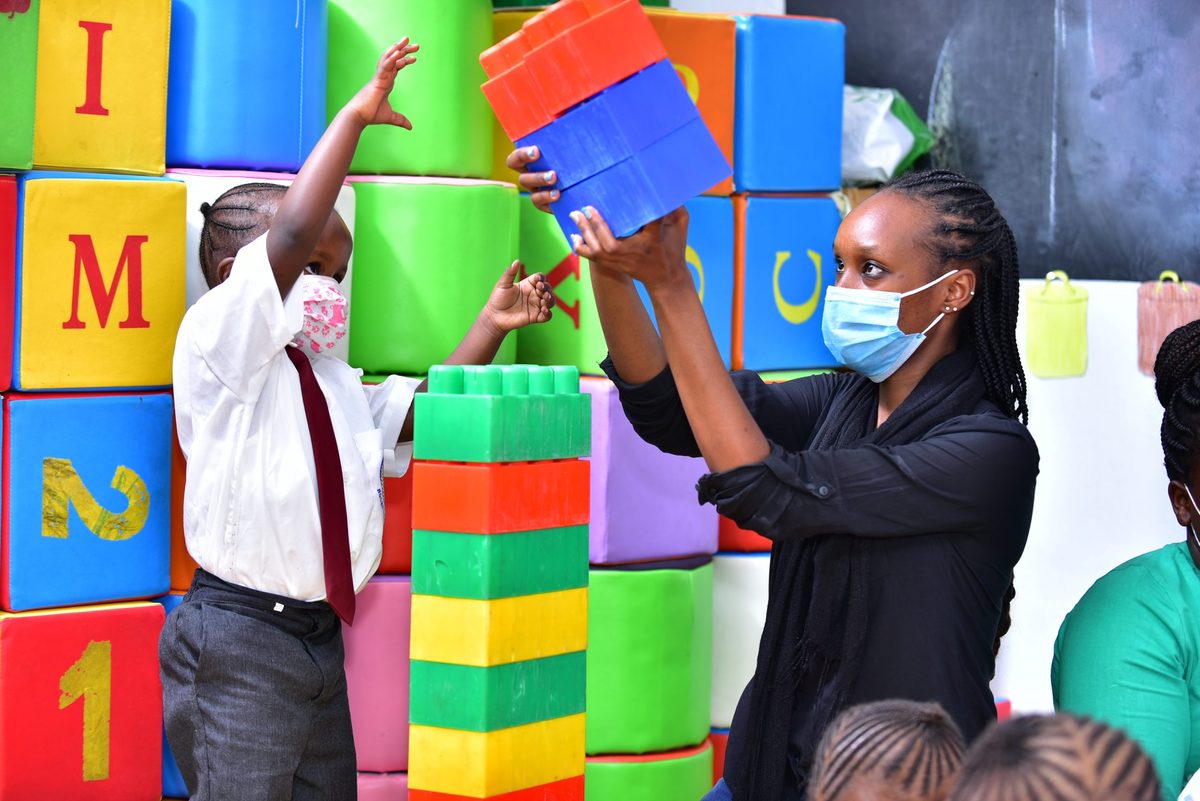 A kindergarten teacher lifting a giant building block with a little girl in a tie reaching up